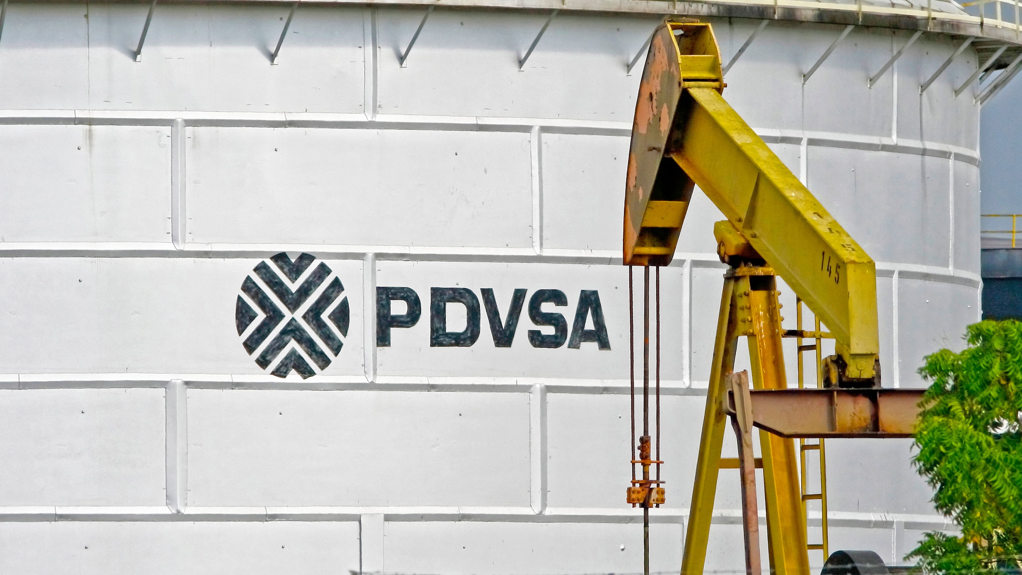PDVSA logo on a crude storage tank, with a yellow pump jack rig in front of it.