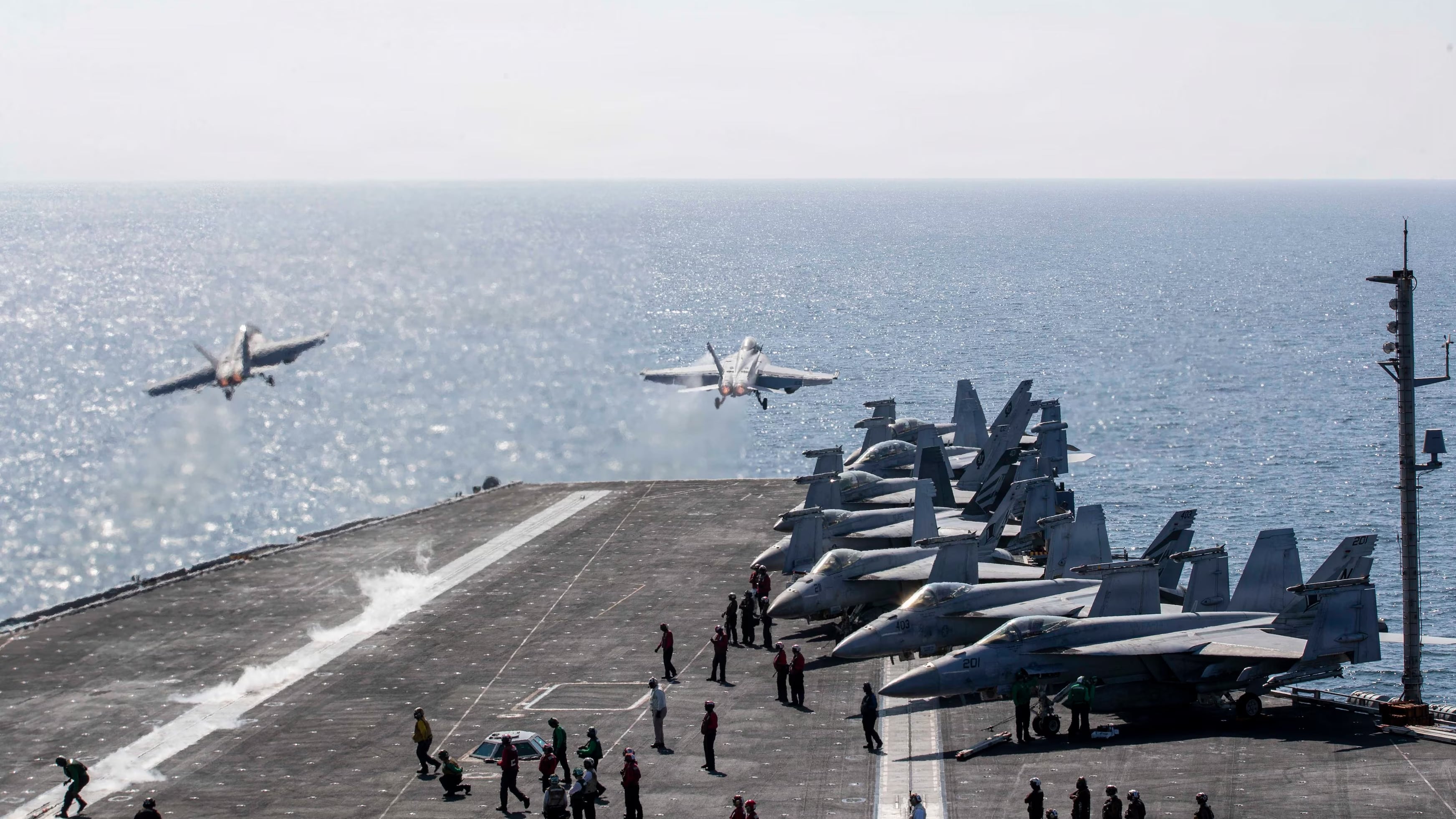 Two fighter jets take off from the deck of an aircraft carrier.